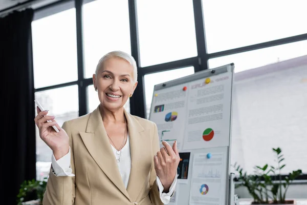 Glücklicher Banker mit Handy, der in die Kamera in der Nähe von verschwommenem Flipchart blickt — Stockfoto