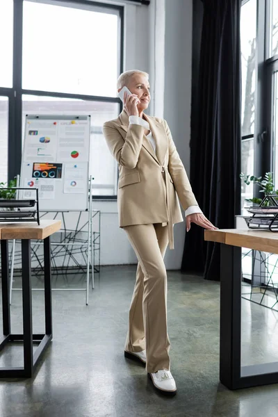 Full length of elegant banker talking on smartphone near flip chart with graphs on background — Stock Photo