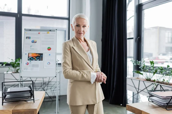 Positive economist smiling at camera near flip chart on blurred background — стоковое фото