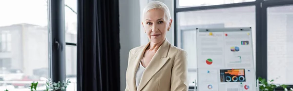 Senior banker smiling at camera near flip chart with graphs on blurred background, banner — Foto stock