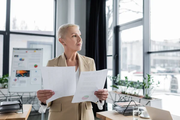 Senior economist looking away while standing with documents in office — Stock Photo