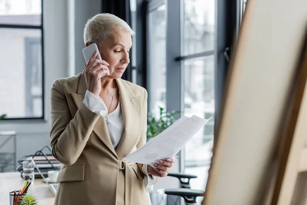 Femme d'affaires aux cheveux gris court tenant des papiers et appelant sur téléphone portable au premier plan flou — Photo de stock