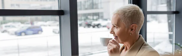 Smiling senior businesswoman looking away while drinking coffee near office windows, banner — Stock Photo