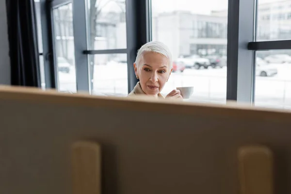 Senior banker with short hair holding coffee cup near blurred flip chart — Stock Photo