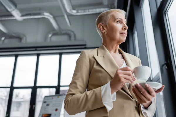 Low angle view of senior banker looking away while standing with coffee cup in office — Photo de stock