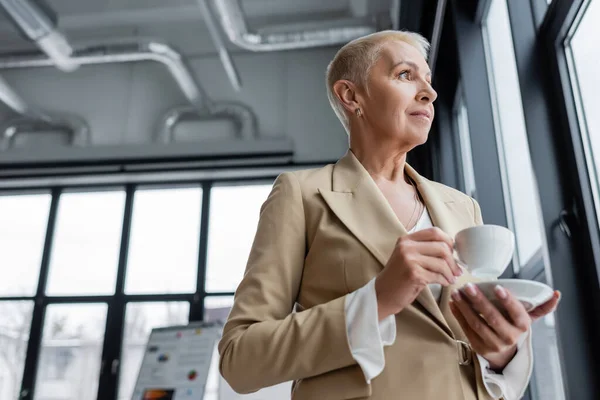 Vue à angle bas du banquier senior regardant loin tout en se tenant debout avec tasse de café dans le bureau — Photo de stock