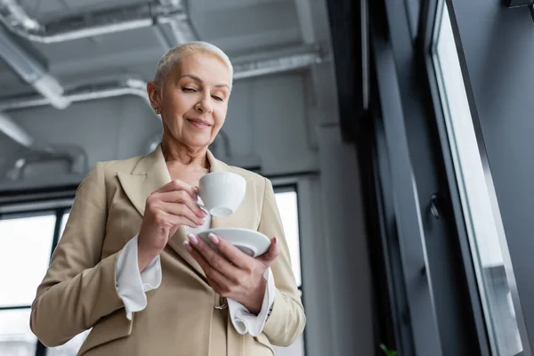 Низький кут зору позитивної старшої бізнес-леді тримає чашку кави в офісі — стокове фото