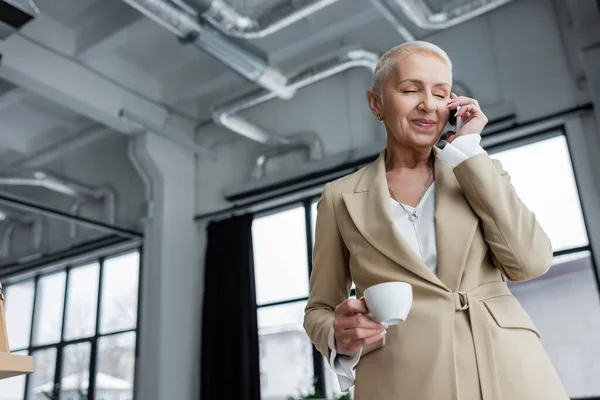 Banchiere sorridente con gli occhi chiusi che parla sullo smartphone mentre tiene in mano una tazza di caffè — Foto stock