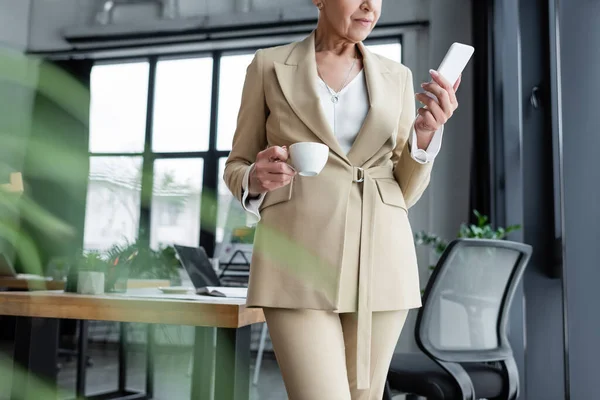 Частичный взгляд на стильную деловую женщину, стоящую со смартфоном и чашкой кофе в офисе — стоковое фото