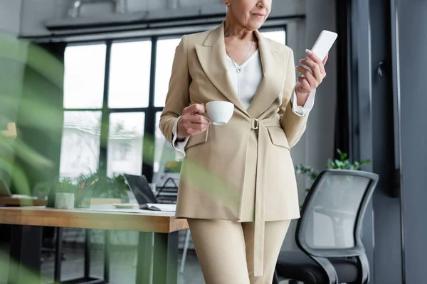 Partial view of stylish businesswoman standing with smartphone and coffee cup in office — Stock Photo