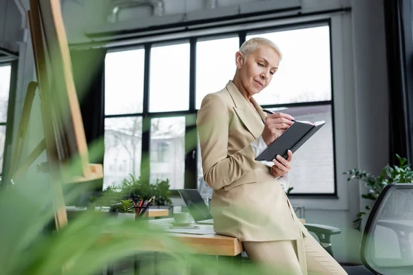 Chefökonom schreibt in Notizbuch, während er sich auf Schreibtisch im verschwommenen Vordergrund lehnt — Stockfoto