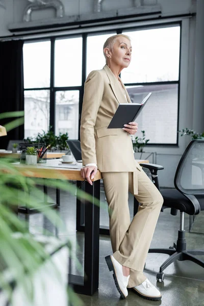 Full length view of stylish banker standing with notebook near desk and looking away — Stock Photo