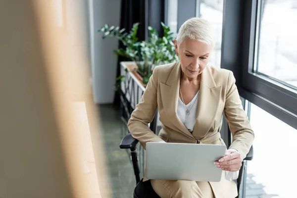 Banquier senior utilisant un ordinateur portable au bureau tout en étant assis sur le premier plan flou — Photo de stock