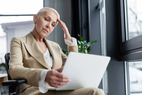 Senior stylish economist looking at laptop and touching forehead — Stock Photo