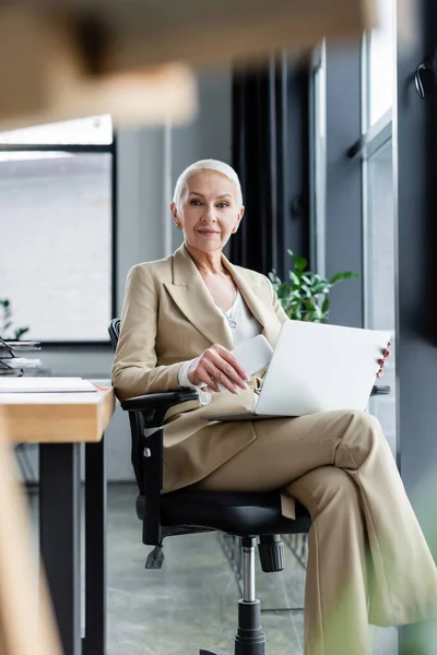 Successful banker smiling at camera while sitting with laptop and smartphone on blurred foreground — Stock Photo