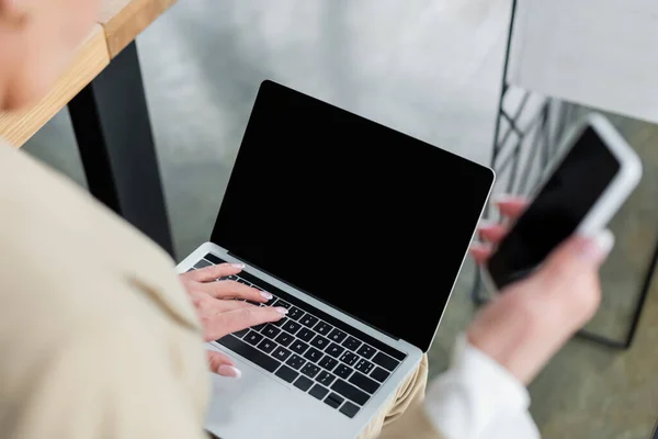 Cropped view of banker with blurred smartphone typing on laptop with blank screen — Fotografia de Stock