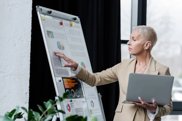 Grey haired and stylish economist with laptop touching graphs on flip chart — Stock Photo