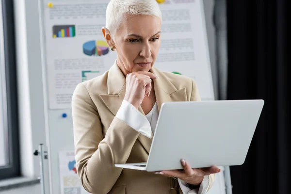 Thoughtful banker standing with laptop near flip chart with infographics on blurred background — Stock Photo