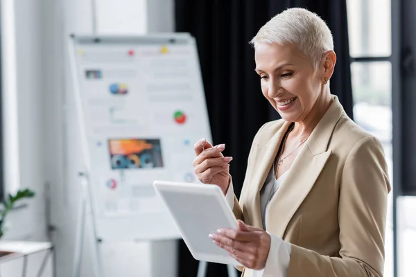 Smiling banker looking at digital tablet near blurred flip chart with infographics — Stock Photo