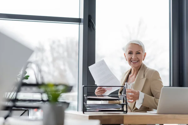Happy senior economist holding water and documents while looking at camera near laptop — Foto stock
