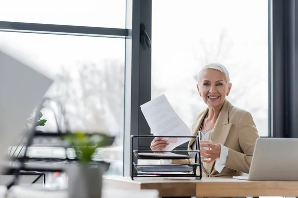 Felice economista senior in possesso di acqua e documenti, mentre guardando la fotocamera vicino al computer portatile — Foto stock