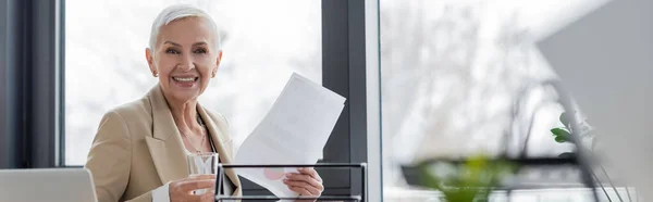 Stylish banker with glass of water and document smiling at camera in office, banner — Stock Photo