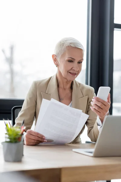 Smiling banker holding documents during video call on smartphone at workplace — Stock Photo