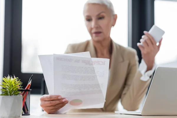 Blurred banker holding papers and smartphone near laptop on desk — Stock Photo