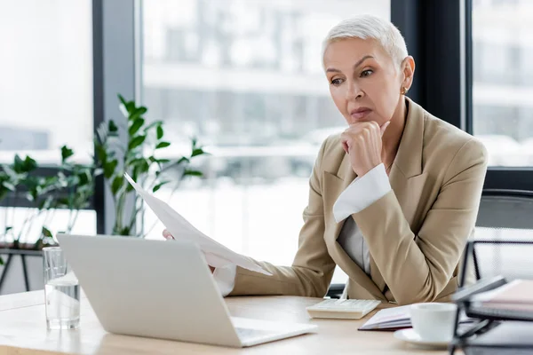 Anziano finanziere in possesso di documenti mentre seduto vicino al computer portatile — Foto stock