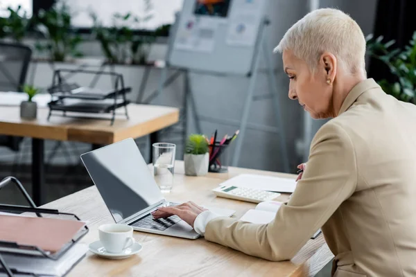 Banquier utilisant un ordinateur portable avec écran vierge près de tasse de café et verre d'eau sur le bureau — Photo de stock