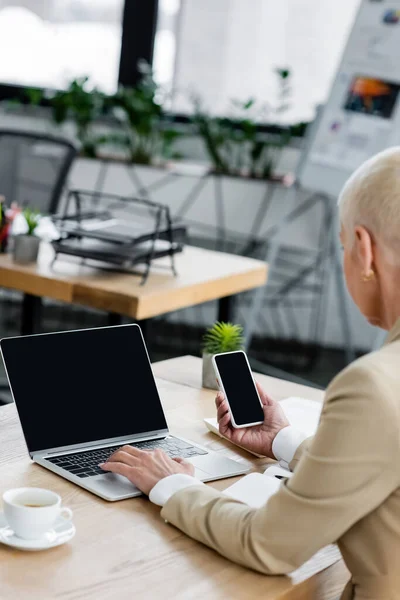 Seniorin hält Smartphone mit leerem Bildschirm beim Tippen auf Laptop — Stockfoto