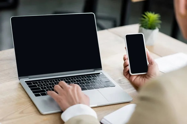 Vista recortada de economista escribiendo en el ordenador portátil y la celebración de teléfono inteligente con pantalla en blanco - foto de stock