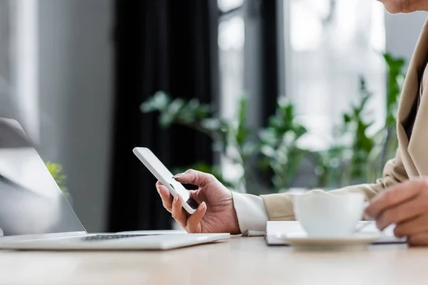 Vue partielle du banquier à l'aide d'un téléphone portable près de l'ordinateur portable et tasse de café floue — Photo de stock