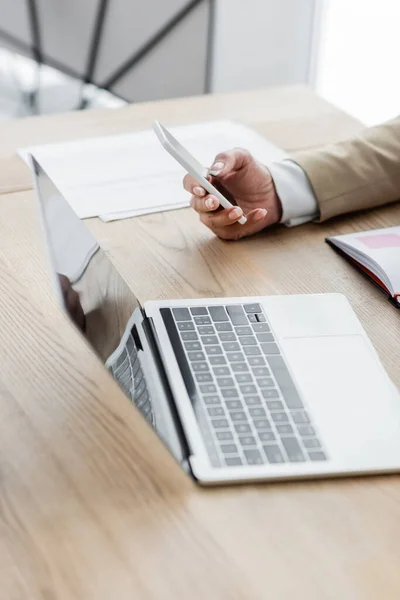 Teilansicht des Ökonomen, der sein Smartphone in der Nähe eines verschwommenen Laptops hält — Stockfoto