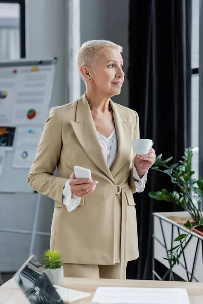 Happy businesswoman with coffee cup and smartphone looking away in office — Stock Photo