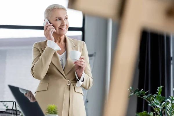 Mujer de negocios sonriente con taza de café hablando en el teléfono móvil en primer plano borrosa - foto de stock