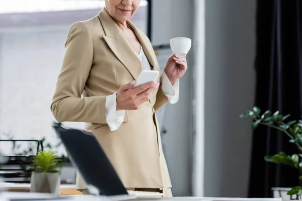 Vista cortada de banqueiro sorridente com smartphone e xícara de café perto de laptop borrado com tela em branco — Fotografia de Stock