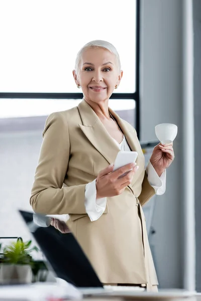 Exitoso banquero sonriendo a la cámara mientras sostiene el teléfono inteligente y la taza de café - foto de stock