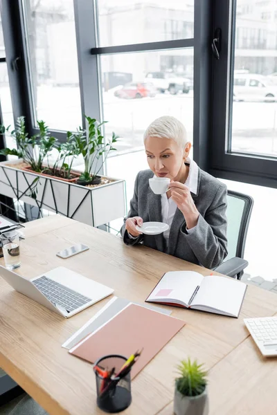 Economista senior beber café en el lugar de trabajo cerca de la computadora portátil y portátil en blanco - foto de stock