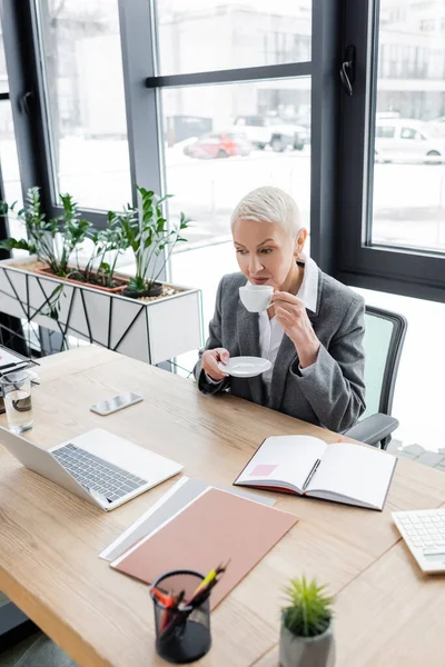 Senior economist drinking coffee at workplace near laptop and blank notebook — стоковое фото