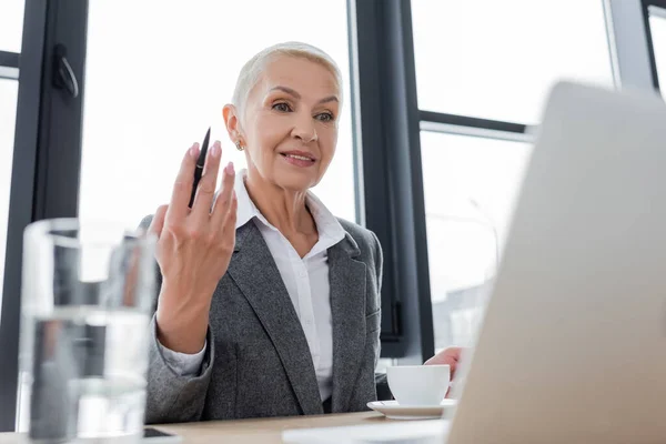 Positive Geschäftsfrau hält Stift während Videochat auf Laptop in der Nähe Kaffeetasse und verschwommenes Glas Wasser — Stockfoto