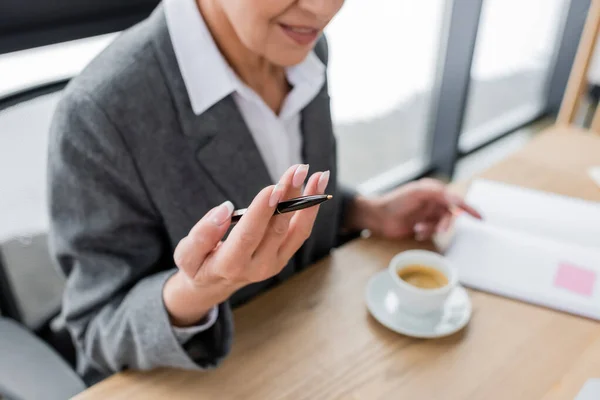 Cropped view of banker with pen sitting at desk with coffee cup in office - foto de stock