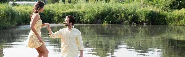 Hombre feliz cogido de la mano de la mujer joven en vestido cerca del lago, bandera - foto de stock