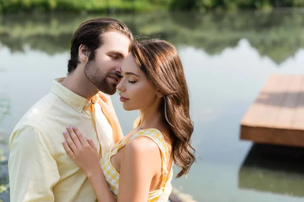 Mujer joven con los ojos cerrados abrazándose con el hombre barbudo cerca del lago - foto de stock