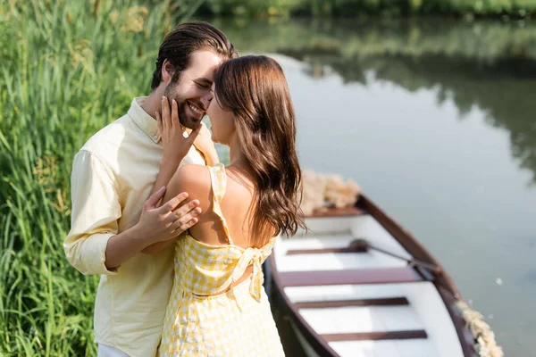 Joven mujer abrazando con feliz barbudo hombre cerca del lago y borrosa barco - foto de stock