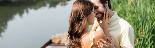 Mujer joven abrazándose con el hombre barbudo feliz cerca del lago, bandera - foto de stock