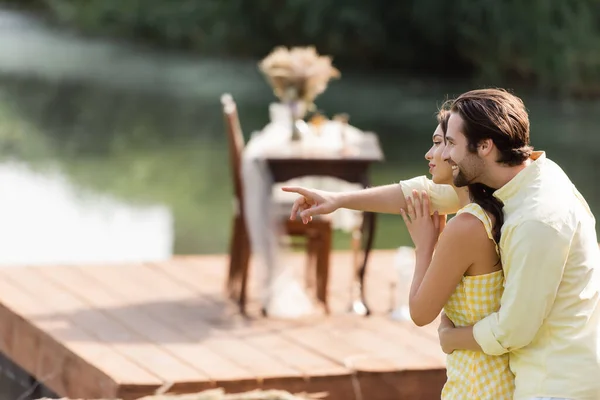 Vista lateral del hombre barbudo feliz abrazando a la novia mientras señala hacia fuera - foto de stock