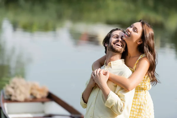 Mujer joven en vestido abrazando feliz novio barbudo cerca del lago - foto de stock