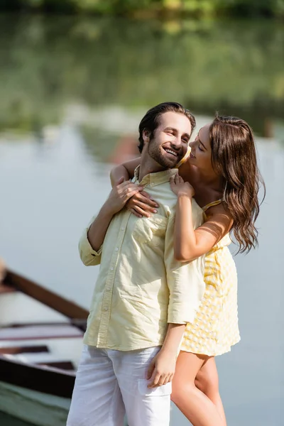 Heureuse jeune femme en robe étreignant copain barbu près du lac — Photo de stock