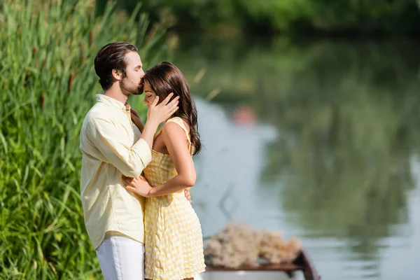 Vue latérale de l'homme barbu embrassant le front de la jeune femme en robe près du lac — Photo de stock